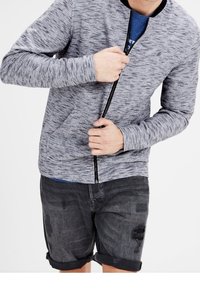 Grå melerad zip-up hoodie med fickor, har en svart krage och ribbade muddar, bärs över en blå skjorta och svarta slitna shorts.