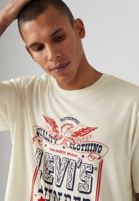 Crème kleurig katoenen T-shirt met een gedetailleerde rode en blauwe grafiek met een adelaar en "Levi’s" branding. Ontspannen ronde halslijn.