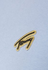 Světl blue bavlněná tkanina s texturovaným povrchem, na které je vyšívané logo "Tommy" ve zlaté a černé barvě v stylizovaném písmu.