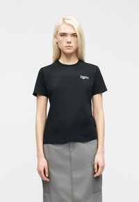 Νεαρό άτομο με μακριά ξανθά μαλλιά φοράει μαύρο κοντομάνικο t-shirt με μικρό λευκό λογότυπο Converse και γκρι φούστα με τσέπες.