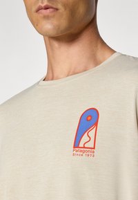 Beżowa koszulka z krótkim rękawem z niebieskim i czerwonym logo Patagonia na klatce piersiowej. Gładka tekstura, okrągły dekolt i standardowy krój.