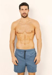 Pánské plavky v modré tkanině. Šortky mají elastický pas s černou šňůrkou a bočními kapsami. Hladká textura.