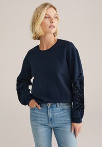 Blonda ženska s kratkimi lasmi, oblečena v mornarsko modro majico s črnimi cvetličnimi motivi na rokavih in svetlo modre kavbojke, pozira z roko v žepu.
