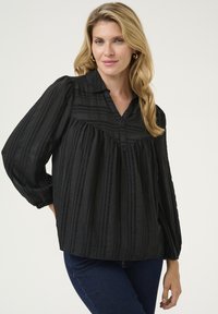 Melna blūze no teksturēta auduma ar vertikālām svītrām, ar apkakles dekoltē un apjomīgām piedurknēm.