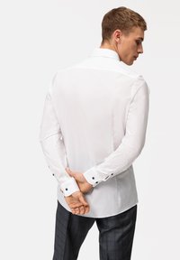 Weißes Hemd mit glatter Textur, langen Ärmeln und schwarzen Knopfakzenten an den Manschetten, getragen mit grauen karierten Hosen.