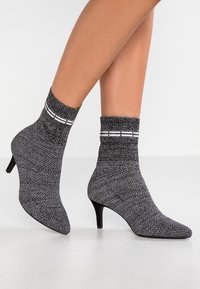 Bottine à chaussette grise texturée avec un bout pointu et un talon aiguille noir. Présente un haut de chaussette côtelé avec des rayures blanches.