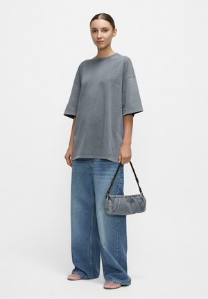 Femme portant un t-shirt gris surdimensionné et un jean bleu à larges jambes, tenant un petit sac à main gris froncé avec une sangle noire.