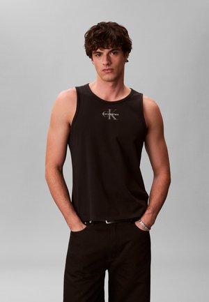 Junger Mann mit lockigem Haar, der ein schwarzes ärmelloses Calvin Klein Top und schwarze Hosen trägt, mit den Händen in den Taschen vor einem grauen Hintergrund.