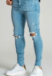 Piernas vestidas con jeans skinny desgastados de color azul claro y zapatillas blancas con detalles dorados y negros, de pie contra un fondo sencillo.