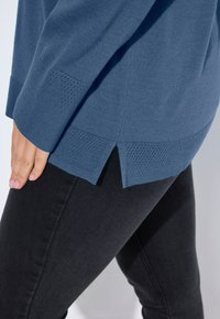 Sininen neulepaita, jossa on rento fit, teksturoidut yksityiskohdat helmassa ja sivuhalkiot, yhdistettynä mustiin farkkuihin.