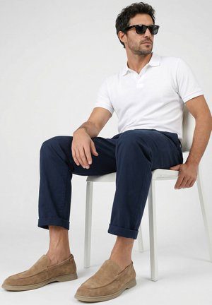 Mann trägt schwarze Sonnenbrille, weißes Poloshirt, dunkelblaue Hose und beige Slipper, sitzt auf einem weißen Stuhl vor schlichtem Hintergrund.