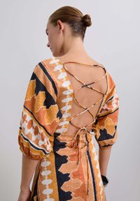 Vue arrière d'une robe à motifs avec des couleurs orange, noire et blanche. La robe présente un dos en V profond avec des bretelles croisées et des manches élastiques.