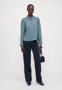 Soft blue long-sleeve blouse with a ruffled round neckline and keyhole detail, paired with dark navy wide-leg trousers and a small black handbag.