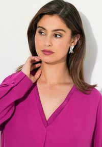 Blusa fucsia con scollatura a V e polsini bottone, realizzata in tessuto morbido. Orecchini decorativi in argento e capelli naturali che incorniciano il viso.