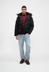 Juoda pūkinė striukė su aukštu apykakle, derinama su bordo spalvos marškiniais ir šviesiais mėlynais laisvais džinsais. Aprangą užbaigia rudos spalvos batai.