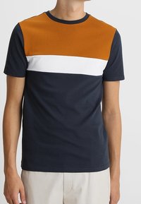 Pánské tričko z námořnického modrého bavlněného materiálu s oranžovým horním panelem, s bílými a oranžovými vodorovnými pruhy přes hrudník. Krátké rukávy.