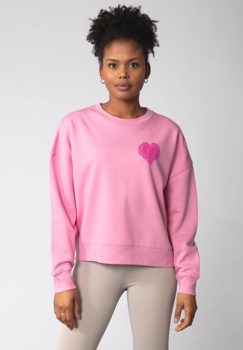Rosa Sweatshirt mit weichem Stoff, übergroßer Passform und Herzdesign auf der Brust, getragen mit gerippten hellen Hosen.