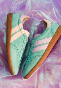 Paire de baskets en daim turquoise avec des bandes blanches et des semelles en caoutchouc marron sur un fond de tissu rose irisé.