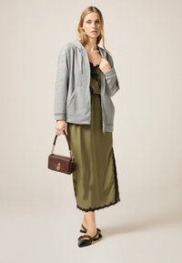 Felpa zip grigia chiara con decorazioni, indossata sopra un vestito di satin verde oliva con bordo in pizzo nero, abbinata a ballerine nere e una borsa marrone.