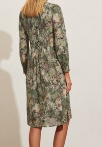 Kwiatowa sukienka w zielonych odcieniach, z marszczonym tyłem, długimi rękawami i bocznymi rozcięciami. Tkanina ma lekką, teksturowaną fakturę.