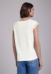 Top blanco de manga corta con bordado a ganchillo en las mangas, con un corte relajado y una tela suave y texturizada, combinado con pantalones de mezclilla azul claro.