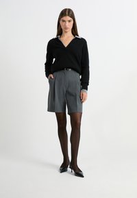 Schwarzer V-Ausschnitt-Pullover mit einem hellgrauen Kragen, kombiniert mit grauen maßgeschneiderten Shorts und schwarzen schnittigen Pumps. Model trägt transparente schwarze Strumpfhosen.