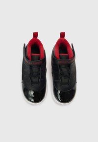 Μαύρα αθλητικά παπούτσια με γυαλιστερό φινίρισμα από δερματίνη, κόκκινη ύφανση στο εσωτερικό, ελαστικά κορδόνια και θηλιά στο πίσω μέρος της φτέρνας.