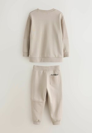 Vaatteet setti, jossa on beige collegepaita ja collegehousut. Collegepaidassa on pyöreä kaula-aukko ja ribbineulehihat; housuissa on sivutasku logolla ja teksturoidut kangasdetaljit.