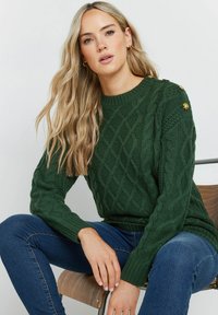 Zielony sweter robiony na drutach z warkoczowym wzorem, okrągłym dekoltem i guzikami na ramionach. W parze z niebieskimi dżinsami, siedzący na drewnianym krześle.