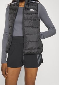 Svart vadderad väst med hög krage, featuring vit Adidas-logga och tre vita ränder på axlarna, kombinerad med svarta shorts.