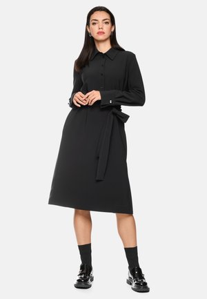 Woman standing wearing a long-sleeve black shirt dress with a waist tie and black ankle socks paired with shiny black shoes.