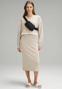 Sweatshirt beige à épaules tombantes, associé à une jupe midi beige ajustée. Un sac banane noir est porté en bandoulière, avec des sandales blanches à enfiler.