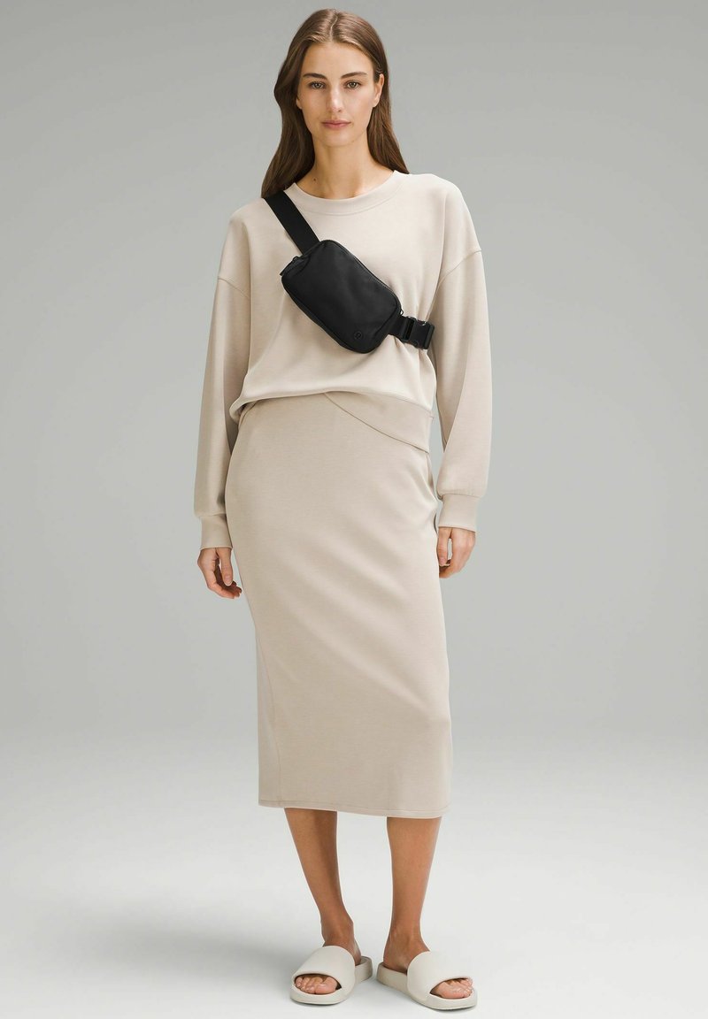 Sweatshirt beige à épaules tombantes, associé à une jupe midi beige ajustée. Un sac banane noir est porté en bandoulière, avec des sandales blanches à enfiler.