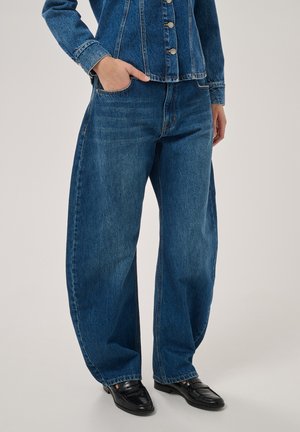 Personne portant un jean denim bleu ample, une veste denim assortie et des mocassins en cuir noir, debout avec une main dans la poche.