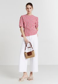 Donna che indossa una camicia a quadri rossi e bianchi e pantaloni bianchi a gamba larga, tiene in mano una piccola borsa intrecciata con dettagli in pelle marrone.