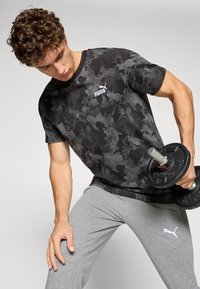 Zwarte trainings-T-shirt met camouflagepatroon, ronde hals, korte mouwen en wit Puma-logo, gecombineerd met grijze joggingbroek en een gewicht.
