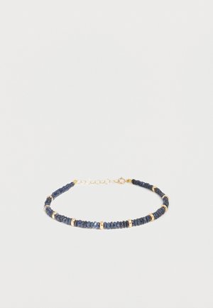 Bracelet délicat avec de petites perles bleu foncé entrecoupées d’anneaux dorés et un fermoir chaîne doré sur fond uni.