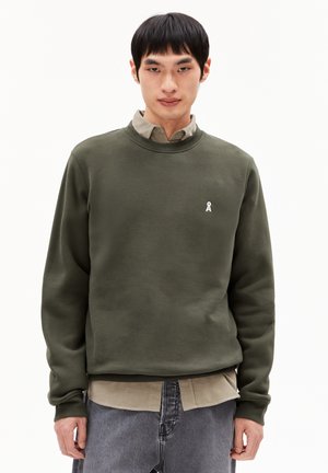 Sweatshirt vert olive en tissu doux, avec un col rond et un petit logo blanc. Le sweatshirt est superposé à une chemise beige.
