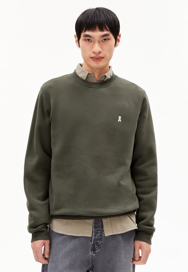 Olijfgroene sweatshirt gemaakt van zachte stof, met een ronde halslijn en een klein logo in het wit. De sweatshirt is gelaagd over een beige shirt.