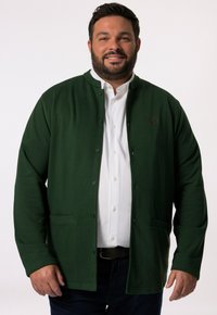 Grön knäppjacka med rundad krage, tillverkad av mjukt tyg. Har sidofickor och en diskret logotyp på vänster bröstområde.