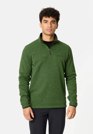 Jonge man draagt een groene fleece trui met kwart rits en zwarte broek, staand tegen een effen lichte achtergrond.