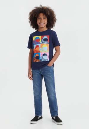 Göndör hajú fiú sötétkék pólót visel, amelyen hat színes űrhajós sisak mintája látható, valamint kék farmert és fekete bebújós cipőket.