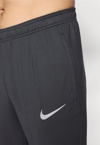 Sorte træningsbukser med elastisk talje, sidelommer og et lysegrå broderet Nike-logo på venstre ben. Glat tekstur.