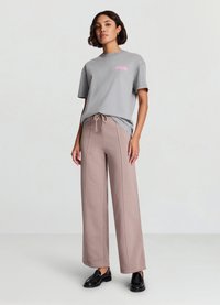 T-shirt in cotone grigio con grafica rosa, abbinata a pantaloni sportivi beige a gamba larga con coulisse e cuciture visibili. Scarpe nere.