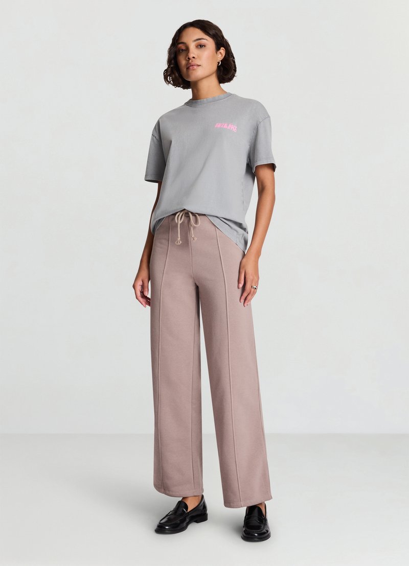 T-shirt in cotone grigio con grafica rosa, abbinata a pantaloni sportivi beige a gamba larga con coulisse e cuciture visibili. Scarpe nere.