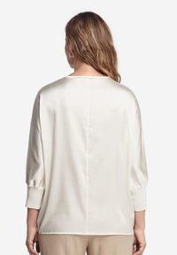 Žena se světlými hnědými vlnitými vlasy, oblečená v volné bílé saténové blouse s manžetovými tříčtvrtinovými rukávy, viděná zezadu.