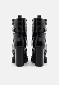 Bottes courtes en cuir verni noir avec un talon bloc haut, des boucles décoratives et un détail zippé sur les côtés. Finition lisse et brillante.