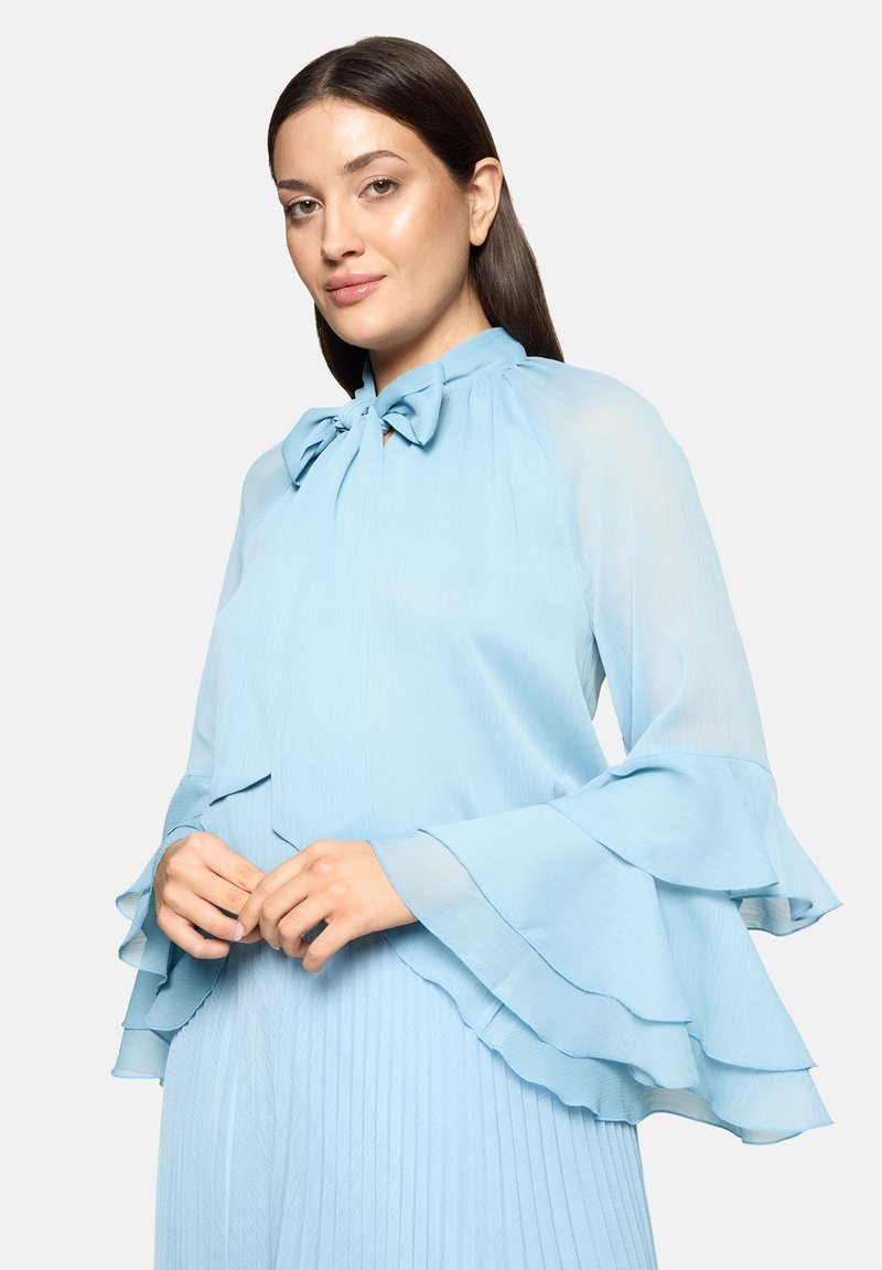 Blouse bleu clair en chiffon avec un col montant et un détail de nœud, dotée de manches à volants superposés et de plis verticaux le long du corps.