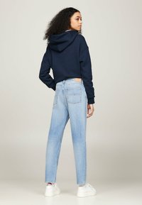 Mujer con cabello rizado que lleva una sudadera azul marino, jeans de tiro alto azul claro, calcetines blancos y zapatillas blancas, de pie contra un fondo sencillo.