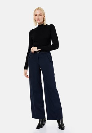 Femme blonde portant un col roulé noir, un pantalon large bleu marine avec ceinture, des bottes noires pointues, debout avec une main sur la hanche sur fond blanc.